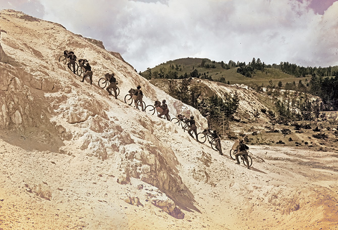 Enlargeable sepia tone image of soldiers walking their bikes up a mountain