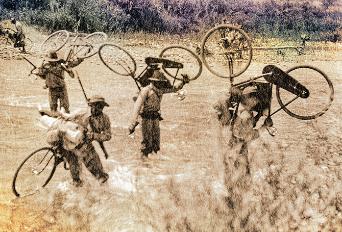 Sepia tone image of soldiers carrying bikes through a field